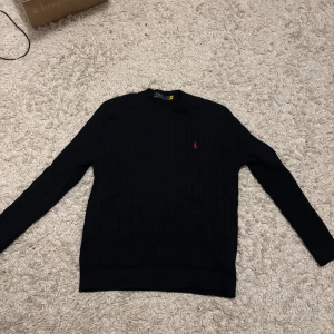 Mörkblå stickad tröja Polo Ralph Lauren - Kabelstickad mörkblå tröja från Polo Ralph Lauren med klassisk röd broderad logga på bröstet. Tröjan har rund halsringning, ribbade muddar och långa ärmar. Perfekt för lager-på-lager och snygg till jeans eller kjol.