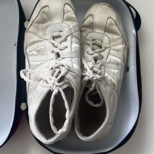 Vita cheerleading skor från Nfinity - Säljer ett par vita cheerleading sneakers från Nfinity. Skorna har snörning, rund tå och är tillverkade i syntetmaterial med meshdetaljer för extra andningsförmåga. Kommer med original röd och vit hård väska med Nfinity-logga. Perfekta för träning och cheer.