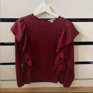Vinröd volangblus - Säljer en vinröd tröja från H&M med volangdetaljer. Tröjan har en rund halsringning med en knapp bak.