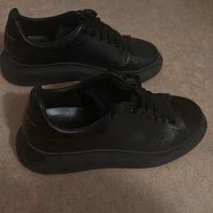 Svarta sneakers från Alexander McQueen - Säljer ett par helsvarta sneakers från Alexander McQueen med chunky sula och klassisk snörning. Skorna är tillverkade i skinn och har diskret logga i guld på hälen. Perfekta för dig som gillar stilrena och trendiga skor med lite extra edge. Kan gå ner i pris vid snabbaffär