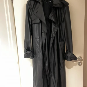 Svart trenchcoat i skinn från Zara - Lång svart trenchcoat från Zara i snyggt skinnmaterial. Jackan har breda slag, bälte vid ärmsluten och stora fickor framtill. Perfekt för dig som vill ha en edgy och stilren look. Oversized passform och klassisk design.