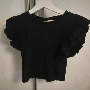 Svart croppad topp med volangärm - Tidigare trendig snygg svart croppad topp med korta volangärmar som ger en cool och trendig look. Perfekt att matcha med höga jeans eller kjol. Mjuk och skön bomullskänsla, enkel att styla för olika tillfällen.