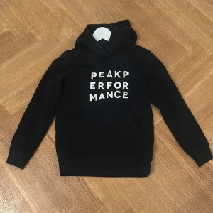 Svart hoodie från Peak Performance - Svart hoodie från Peak Performance med vit text på bröstet. Klassisk känguruficka framtill och huva. Perfekt för en chill och sportig stil. Mjuk insida och ribbade muddar vid ärmslut och nederkant.