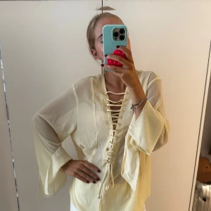 Ljusgul blus med snörning - Säljer en ljusgul blus i transparent, lätt material med vida ärmar och snygg snörning framtill. Blusen har en avslappnad passform och v-ringning, perfekt för dig som gillar boho-vibes och unika detaljer.