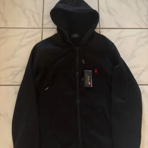 Svart hoodie och byxor från Polo Ralph Lauren - Svart hoodie och byxor från Polo Ralph. Klassisk design med röd broderad logga på bröstet och snörning i huvan. Tillverkad i mjuk bomull, perfekt för chill dagar. Fickor framtill för extra bekvämlighet.