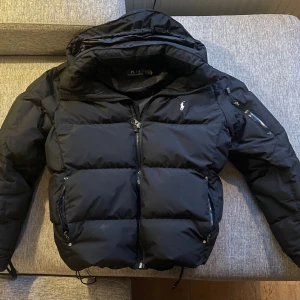 Svart pufferjacka från Polo Ralph Lauren - Säljer en svart pufferjacka från Polo Ralph Lauren med klassisk logga på bröstet. Jackan har huva, dragkedja framtill och praktiska fickor med dragkedja. Perfekt för kalla dagar och riktigt snygg till jeans eller sweatpants. Mycket bra skick efter lite användning, inga täcken på användning. Säljer eftersom jag köpt en annan.