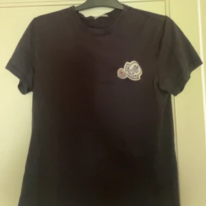 Svart Moncler t-shirt med patch - Svart t-shirt från Moncler med rund hals och korta ärmar. På bröstet sitter en stor broderad Moncler-patch i vitt, blått och rött. T-shirten är i klassisk modell och tillverkad i mjuk bomull som känns skön mot huden.