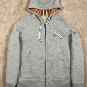Grå hoodie från Burberry med dragkedja - Snygg grå hoodie från Burberry med klassiskt rutigt foder i huvan och dragkedja framtill. Tröjan har snörning i huvan, två fickor och diskret Burberry-logga på bröstet. Perfekt för en chill och stilren look.