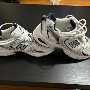 Vita New Balance 530 sneakers - Snygga New Balance 530 sneakers i vit och marinblå färg med mesh och syntetmaterial. Klassisk rund tå, platt sula och snörning framtill. Ikonisk N-logga på sidan och detaljer i silver. Perfekta för dig som gillar sportig stil och komfort.