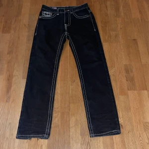 Svarta True Religion jeans med kontrastsömmar - Säljer ett par svarta jeans från True Religion med tydliga vita kontrastsömmar och ikoniska fickdetaljer. Modellen har raka ben och klassisk femficksdesign. Jeansen är tillverkade i USA och har snygga metallknappar och logotyp på bakfickan.