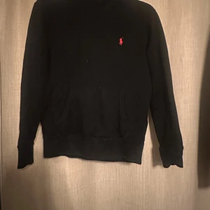 Svart hoodie från Polo Ralph Lauren - Svart hoodie från Polo Ralph Lauren med klassisk röd logga broderad på bröstet. Tröjan har huva, känguruficka och långa ärmar. Perfekt för dig som gillar stilren och enkel design med en touch av lyx.