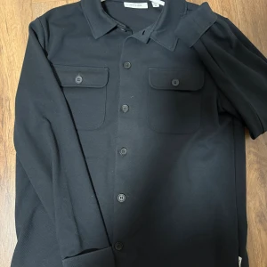 Mörkblå overshirt från Only & Sons - Stilren svart overshirt från Only & Sons med klassisk krage, två bröstfickor med lock och knappar framtill. Skjortan har lång ärm och är tillverkad i ett mjukt material som känns bekvämt. Perfekt att bära öppen eller stängd för en cool look.