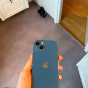 iPhone 13 Blå - Snygg blå iPhone 13 med dubbla kameror. Telefonen har synliga repor på både baksida och skärm, men är i fullt fungerande skick. Perfekt för dig som vill ha en kraftfull iPhone till ett bra pris. Levereras utan tillbehör.