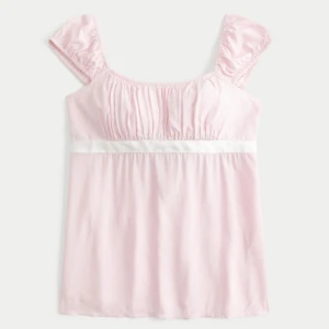 Ljusrosa blus med rosett och puffärm - Supersöt ljusrosa blus med små vita prickar, puffärmar och en vit rosett som knyts i ryggen. Blusen har en mjuk, rynkad överdel och ett band under bysten som ger en feminin siluett. Perfekt till jeans eller kjol för en gullig look.