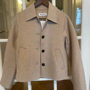 Beige ulljacka från Wera, strl 34 - Snygg beige jacka (Muscat) från Wera i ullmix med klassisk krage och raka fickor framtill. Jackan har en stilren look med knappar och en rak, lite kortare passform. Helt ny med lapparna kvar! 