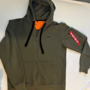 Olivgrön hoodie från Alpha Industries - Säljer en olivgrön hoodie från Alpha Industries med dragkedja, huva och svarta snören. Tröjan har en ikonisk röd 'Remove Before Flight'-tag på ärmen, orange insida i huvan och praktiska fickor. Perfekt för dig som gillar streetwear och snygga detaljer.