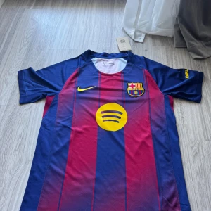 FC Barcelona hemmaställ 25/26 (Rabatt) - Barças officiella matchtröja från Nike i Dri-FIT-material. Klassiska blå och vinröda ränder, klubbmärke på bröstet, gul Nike-logga och Spotify-sponsor framtill. UNHCR-tryck på ryggen. Perfekt för dig som älskar fotboll och vill visa ditt stöd för Barca.