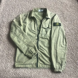 Grön overshirt från Stone Island - Snygg ljusgrön overshirt från Stone Island med klassisk patch på ärmen. Jackan har dragkedja framtill, bröstficka med knapp och elastiska muddar vid ärmslut. Perfekt för lager-på-lager. Passar dig mellan 160~175. Fixar hål i armhålan DÄRAV priset! Nypris: 3800kr MITT pris: 999kr (kan gå ner vid snabb affär) Har du några frågor kring jackan är det bara att hugga till! MVH. SWISSWEAR🇨🇭🍾🏔️