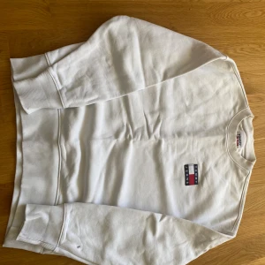 Vit sweatshirt från Tommy Jeans - Vit sweatshirt från Tommy Jeans med klassisk logga broderad på bröstet. Tröjan har rund hals, långa ärmar och ribbade muddar vid ärmslut och nederkant. Perfekt för en clean och stilren look.