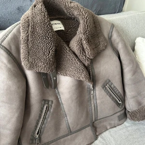 Brun teddyjacka från Zara med dragkedjor - Säljer en brun teddyjacka från Zara med stora mysiga krage, detaljer i skinnimitation och coola dragkedjor på fickorna. Jackan har breda slag, markerade sömmar och en oversized vibe. Perfekt för kalla dagar när du vill vara både varm och snygg.