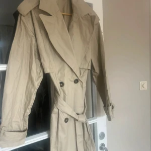Beige trenchcoat med bälte - Stilren beige trenchcoat med klassisk krage, dubbelknäppning och bälte i midjan. Jackan har långa ärmar med knäppning vid ärmslut och stora slag. Perfekt för dig som vill ha en tidlös och snygg look.