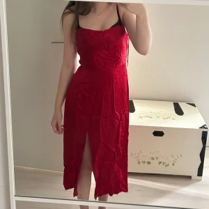 Röd midiklänning med slits från Zara - Röd midiklänning från Zara Basic med smala axelband, knyt i ryggen och en snygg slits framtill. Klänningen har en figurnära passform och är tillverkad i ett glansigt, lätt material som ger en elegant känsla.