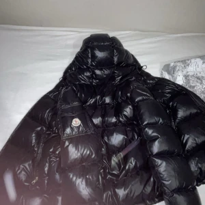 Svart pufferjacka från Moncler Äkta XXL Maya - Säljer en svart, glansig pufferjacka från Moncler med huva och dragkedja. Jackan har en klassisk Moncler-logga på bröstfickan och stora quiltade partier. Perfekt för kalla dagar och riktigt snygg streetstil.