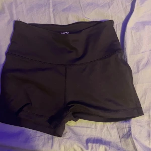 Svarta högmidjade träningsshorts Craft XS - Snygga svarta träningsshorts från Craft i storlek XS. De är högmidjade med bred linning och har en tight passform som sitter skönt under träning. Materialet är mjukt och stretchigt polyester, perfekt för gymmet eller löprundan. Snygga sömmar ger en sportig vibe.