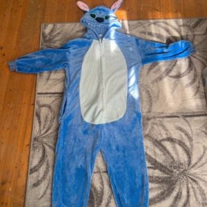 Blå Stitch onesie med huva - Supermysig blå Stitch onesie med huva, stora broderade ögon, rosa öron och vita tänder. Dragkedja framtill och mjukt fleece-material. Perfekt för dig som vill chilla hemma eller sticka ut på maskerad. Söt detalj med liten svans bak.