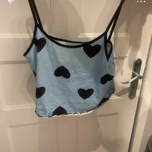 Blå linnetopp med svarta hjärtan - Supersöt ljusblå linnetopp med svarta hjärtan och svarta kantband. Toppen är ärmlös med tunna axelband och har en lätt croppad passform. Tillverkad i mjuk polyesterblandning som känns skön mot huden. Perfekt för dig som vill sticka ut med ett gulligt mönster.