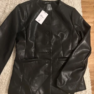 Svart skinnjacka från Gina Tricot - Snygg svart skinnjacka från Gina Tricot med rund hals och knäppning med fem knappar framtill. Jackan har två fickor fram och en stilren, figurnära passform. Perfekt att styla med jeans eller kjol för en cool look.