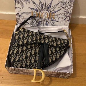 Svart o beige mönstrad väska - Ikonisk väska med svart och beige monogrammönster, guldfärgade metalldetaljer och stort D-hänge. Väskan har en unik sadelform och är tillverkad i canvas med detaljer i skinn. Långt band och låda tillkommer.🫶🏻