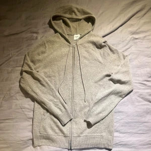 Beige cashmere hoodie från John Henric - Mjuk och lyxig beige hoodie i 100% kashmir från John Henric. Tröjan har dragkedja framtill, huva med snörning och två fickor. Perfekt för dig som vill ha en stilren och bekväm look med premiumkänsla.