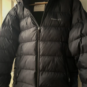 Marinblå pufferjacka från GANT - Marinblå pufferjacka från GANT med huva och dragkedja framtill. Jackan har quiltad design, två sidofickor med dragkedja och GANT-logga på bröstet. Perfekt för kalla dagar och riktigt skön att bära. Köpt för 3000kr, säljer för 2500kr! Använd 1 gång.