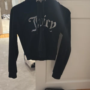 Svart Juicy Couture hoodie med strass - Svart hoodie från Juicy Couture i mjuk sammet med glittrande strassdetaljer på bröstet där det står 'Juicy'. Tröjan har huva, snörning och är croppad i modellen. Perfekt för dig som vill ha en ikonisk och trendig look. Sparsamt använd, köpte begagnat för ett år sen❤️ Säljer då jag rensat gaderoben🥰