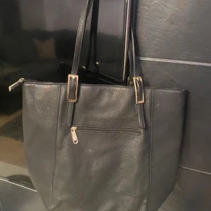 Svart axelväska med gulddetaljer - Snygg svart axelväska från David Jones i skinnimitation med guldfärgade spännen och dragkedja. Väskan har både handtag och avtagbar axelrem, samt dekorativa detaljer längs sidorna. Perfekt storlek för att få plats med allt du behöver.