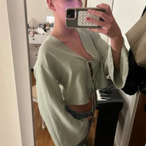 Ljusgrön croppad blus med knyt - Säljer en ljusgrön croppad blus med vida ärmar och tre knyt framtill. Toppen har v-ringning och är tillverkad i ett lätt, flowy material som känns luftigt. Perfekt för dig som gillar en chill och trendig stil.