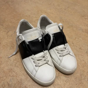 Valentino Garavani vita och svarta sneakers - Snygga Valentino Garavani sneakers i vitt skinn med bred svart läderdetalj över vristen. Klassisk rund tå, vita snören och diskret logga på plösen. Sulan har coola nitar baktill för extra edge. Kommer med originalkartong.