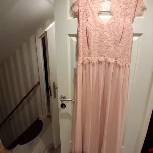 Ljusrosa långklänning med spets - Superfin ljusrosa långklänning med spets upptill och v-ringning. Klänningen har korta ärmar och ett flowy kjolparti i tunt tyg. Spetsdetaljerna ger en romantisk känsla och ryggen har en snygg öppning. Perfekt för speciella tillfällen!