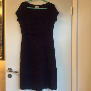 Svart klänning från Rocha John Rocha - Svart klänning med kort ärm från Rocha John Rocha. Klänningen har dekorativa cirkelmönster i flera rader och en rundad urringning. Materialet är mjukt och stretchigt, vilket ger en bekväm passform. Perfekt för dig som gillar unika detaljer och stilren design.