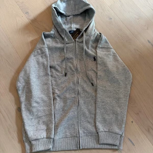 Grå hoodie från Polo Ralph Lauren - Snygg grå hoodie från Polo Ralph Lauren med klassisk logga broderad på bröstet. Tröjan har huva med snörning, dragkedja framtill och ribbade muddar vid ärmslut och nederkant. Perfekt för chill dagar och enkel att matcha med jeans eller joggers.
