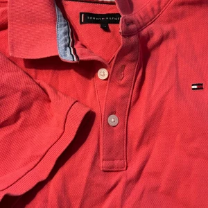 Rosa pikétröja från Tommy Hilfiger - Säljer en snygg rosa pikétröja från Tommy Hilfiger med klassisk krage, knappar framtill och broderad logga på bröstet. Tröjan är i bomull och har korta ärmar med ribbade muddar. Perfekt för en stilren och sportig look.