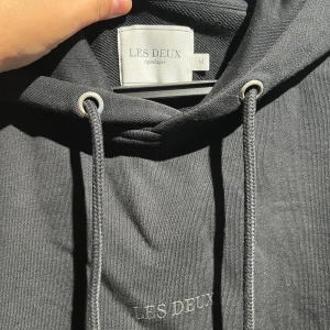Svart hoodie från Les Deux med snörning - Svart hoodie från Les Deux med broderad logga på bröstet, justerbar snörning i huvan och stor känguruficka framtill. Klassisk passform och mjukt material som känns skönt mot huden. Perfekt för dig som gillar stilrena och enkla plagg.