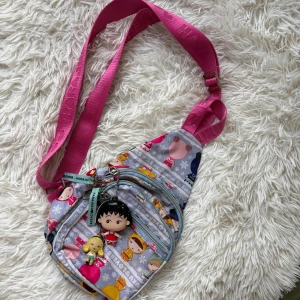 Harajuku väska - Superfin axelväska från Harajuku Lovers med färgglatt figurmönster på ljusblå botten och bred rosa axelrem. Väskan har flera dragkedjefack och söta nyckelringar som detaljer. Perfekt för dig som gillar unika accessoarer med japansk streetstyle-vibe.