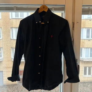 Svart skjorta från Polo Ralph Lauren - Svart skjorta från Polo Ralph Lauren med klassisk button down-krage och röd broderad logga på bröstet. Custom fit-modell med vita knappar och lång ärm. Perfekt för dig som gillar stilrena och tidlösa plagg.