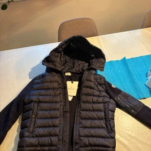 Svart Moncler pufferjacka med huva  - Snygg svart pufferjacka från Moncler med stickade ärmar och vadderad kropp. Jackan har huva, dragkedja framtill, fickor med tryckknappar och en Moncler-logga på ärmen. Perfekt för kyliga dagar och riktigt stilren look. Storlek S säljes endast nå den va för liten för mig