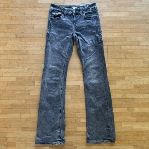 Grå lågmidjade bootcut jeans från Gina Tricot - Snygga grå lågmidjade jeans från Gina Tricot i bootcut-modell. Jeansen har coola detaljer på bakfickorna med broderade mönster och silvriga knappar. Klassisk femficksdesign och bälteshällor. Perfekta för dig som gillar en avslappnad men trendig look.