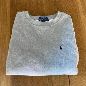 Grå sweatshirt från Polo Ralph Lauren - Klassisk grå sweatshirt från Polo Ralph Lauren med rund halsringning och broderad marinblå logga på bröstet. Tröjan har långa ärmar och ribbade muddar vid ärmslut och nederkant. Perfekt för en avslappnad och stilren look. Den är i fint skick såklart äkta, köpt på NK.