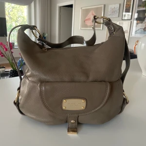 Beige axelväska från Michael Kors - Snygg beige axelväska från Michael Kors i mjukt skinn med guldfärgade metalldetaljer och logoplatta framtill. Väskan har en praktisk dragkedja upptill, justerbar axelrem och en rundad, rymlig form som rymmer allt du behöver till vardagen.