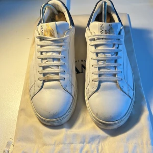 Vita sneakers från Lanvin i skinn - Snygga vita sneakers från Lanvin med blå detalj på hälen och guldfärgad logga på plösen. Skorna är i skinn och har klassisk snörning samt rund tå. Perfekta för dig som gillar stilrena och exklusiva sneakers med en lyxig touch.